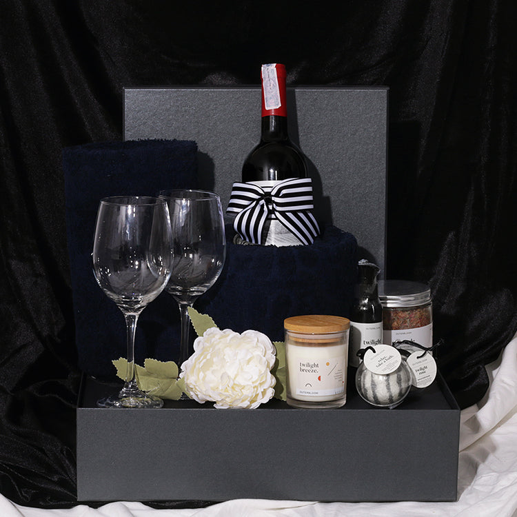 A gift hamper set including a bottle of Rawson's Retreat Shiraz red wine, two wine glasses, a pine and cedarwood soy candle, navy bath towels, a bubble bath, a natural bath bomb, and bath salt, all presented in a black gift box with decorative artificial flowers and a ribbon.