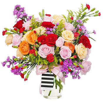 A bountiful and cheerful floral arrangement in a clear glass vase. The bouquet is a vibrant mix of colorful roses in shades of red, peach, cream, and pink, interspersed with small red carnations and purple filler flowers. The arrangement is accented with various green sprigs and foliage, adding texture and depth. A light pink satin ribbon is tied around the vase. This bright and joyful bouquet is a perfect centerpiece for any celebration, expressing happiness and affection.