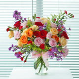 A bountiful and cheerful floral arrangement in a clear glass vase. The bouquet is a vibrant mix of colorful roses in shades of red, peach, cream, and pink, interspersed with small red carnations and purple filler flowers. The arrangement is accented with various green sprigs and foliage, adding texture and depth. A light pink satin ribbon is tied around the vase. This bright and joyful bouquet is a perfect centerpiece for any celebration, expressing happiness and affection.