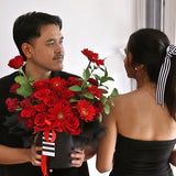 A dramatic and passionate bloom box arrangement overflowing with an assortment of red flowers. It features classic red roses, vibrant red gerberas, and smaller red carnations. The flowers are artfully arranged in a round, matte black box. The arrangement is accented with green leaves and wrapped at the top with ruffled black tissue paper. A striking red satin ribbon and a black-and-white striped ribbon are tied around the box. This bold and romantic gift is perfect for anniversaries and declarations of love