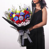 A vibrant and bold bouquet featuring a striking combination of red roses, blue gerberas, and 3 padel/tennis balls. The flowers are arranged with lush green eucalyptus and other filler greens, creating a dynamic contrast. The wrapping is a sophisticated mix of black netted mesh, solid black paper, and a clean white inner layer. The bouquet is tied with a classic black-and-white striped ribbon, adding a modern touch. This powerful arrangement is perfect for celebrating achievements or making a bold state