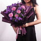 A striking bouquet featuring full pink chrysanthemums and delicate purple asters. It is elegantly wrapped in deep purple paper with a modern black and white striped accent at the base. The arrangement is tied with a pink ribbon bow, creating a full, rounded shape. This sophisticated bouquet is perfect for birthdays, anniversaries, or as a thoughtful thank-you gesture.