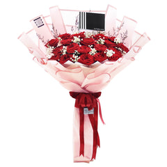 A luxurious and romantic bouquet featuring a generous cluster of approximately 20 red roses. The deep red roses are accented with delicate white baby's breath and thin sprigs of dried filler flowers. The bouquet is wrapped in a soft, semi-transparent pink paper with a subtle striped texture. A double red ribbon is tied into a beautiful bow at the base, and the bouquet includes a stylish black tag. This elegant arrangement is the perfect expression of love and passion.