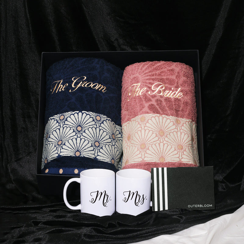 A thoughtful and romantic gift hamper designed for couples. The set is presented in an elegant black box and features two soft towels: a navy blue one and a dusty pink one. Both can be embroidered with custom initials and have a decorative white and pink/blue floral pattern at the bottom. The set also includes two white ceramic mugs with "Mr." and "Mrs." printed on them, along with a small modern card. This beautiful hamper is an ideal gift for weddings, bridal shower, or anniversaries.