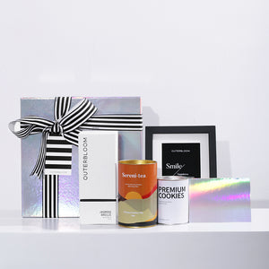A stylish and modern gift hamper featuring a curated selection of items. The contents include: a reed diffuser labeled "JASMINE BREEZE," a tin of premium tea, and white tin of "PREMIUM COOKIES." The hamper also includes a small black picture frame, and a small holographic card. They are all packaged in a large, iridescent holographic box, tied with a bold black-and-white striped ribbon. This gift set is perfect for various occasions, from corporate gift to housewarming.