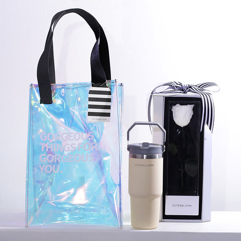 A chic and modern gift set with a white and iridescent theme. The hamper includes a large, transparent holographic tote bag with black handles and the text "GORGEOUS THINGS FOR GORGEOUS YOU." The bag also features a black-and-white striped tag. The set contains a tall, beige tumbler with a grey handle. The set also includes a matching white box that showcases a single, beautifully preserved white rose. This stylish and thoughtful hamper is a perfect gift for someone special.