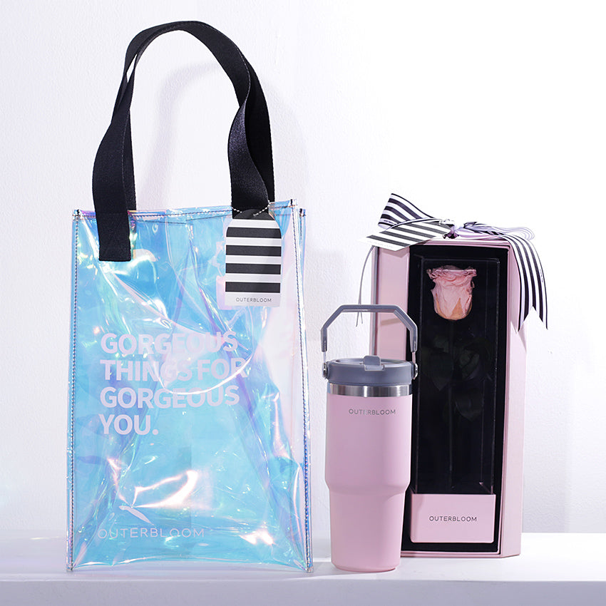 A chic and modern gift set with a pink and iridescent theme. The hamper includes a large, transparent holographic tote bag with black handles and the text "GORGEOUS THINGS FOR GORGEOUS YOU." The bag also features a black-and-white striped tag. The set contains a tall, pink tumbler with a grey handle. The set also includes a matching pink box that showcases a single, beautifully preserved pink rose. This stylish and thoughtful hamper is a perfect gift for someone special.