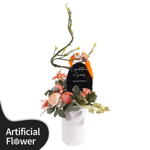 A charming and permanent artificial floral arrangement in a ribbed white ceramic vase. The piece features soft peach and light pink faux peonies and other delicate flowers, accented with realistic green foliage and ivy. An elegant, curving branch extends upwards, where two small orange birds are perched. A black oval plaque is placed in the center, bearing the customizable text "Congratulations on ..". This arrangement is an excellent choice for a housewarming gift or a permanent decor piece.