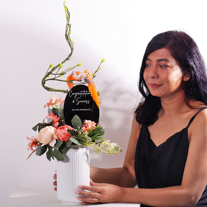 A charming and permanent artificial floral arrangement in a ribbed white ceramic vase. The piece features soft peach and light pink faux peonies and other delicate flowers, accented with realistic green foliage and ivy. An elegant, curving branch extends upwards, where two small orange birds are perched. A black oval plaque is placed in the center, bearing the customizable text "Congratulations on ..". This arrangement is an excellent choice for a housewarming gift or a permanent decor piece.