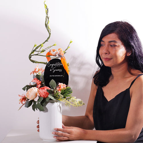 A charming and permanent artificial floral arrangement in a ribbed white ceramic vase. The piece features soft peach and light pink faux peonies and other delicate flowers, accented with realistic green foliage and ivy. An elegant, curving branch extends upwards, where two small orange birds are perched. A black oval plaque is placed in the center, bearing the customizable text "Congratulations on ..". This arrangement is an excellent choice for a housewarming gift or a permanent decor piece.