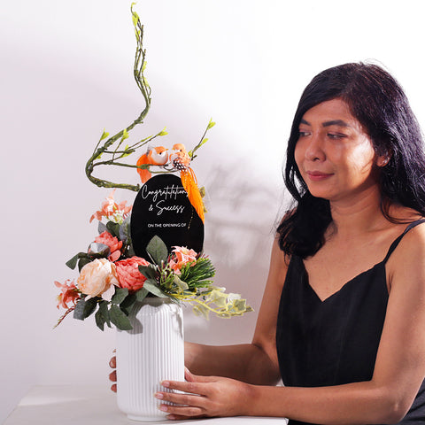 A charming and permanent artificial floral arrangement in a ribbed white ceramic vase. The piece features soft peach and light pink faux peonies and other delicate flowers, accented with realistic green foliage and ivy. An elegant, curving branch extends upwards, where two small orange birds are perched. A black oval plaque is placed in the center, bearing the customizable text "Congratulations on ..". This arrangement is an excellent choice for a housewarming gift or a permanent decor piece.