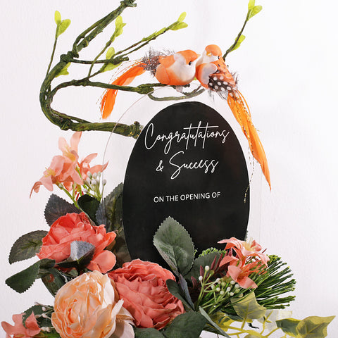 A charming and permanent artificial floral arrangement in a ribbed white ceramic vase. The piece features soft peach and light pink faux peonies and other delicate flowers, accented with realistic green foliage and ivy. An elegant, curving branch extends upwards, where two small orange birds are perched. A black oval plaque is placed in the center, bearing the customizable text "Congratulations on ..". This arrangement is an excellent choice for a housewarming gift or a permanent decor piece.