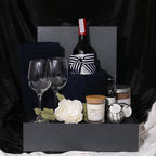 A gift hamper set including a bottle of Rawson's Retreat Shiraz red wine, two wine glasses, a pine and cedarwood soy candle, navy bath towels, a bubble bath, a natural bath bomb, and bath salt, all presented in a black gift box with decorative artificial flowers and a ribbon.