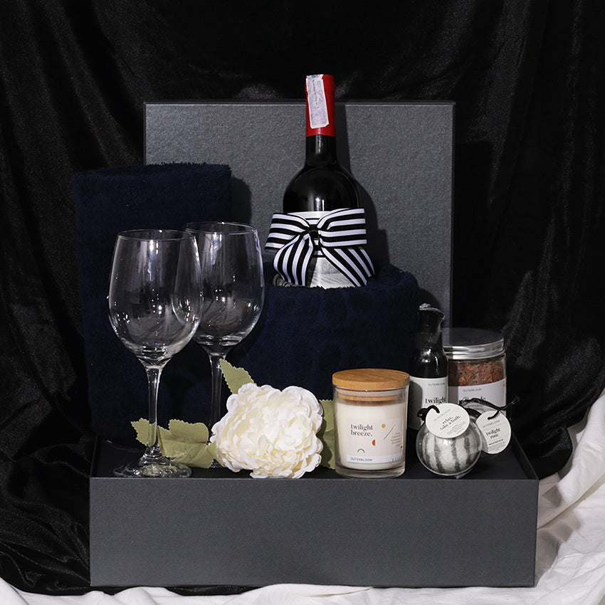 A gift hamper set including a bottle of Rawson's Retreat Shiraz red wine, two wine glasses, a pine and cedarwood soy candle, navy bath towels, a bubble bath, a natural bath bomb, and bath salt, all presented in a black gift box with decorative artificial flowers and a ribbon.