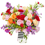 A bountiful and cheerful floral arrangement in a clear glass vase. The bouquet is a vibrant mix of colorful roses in shades of red, peach, cream, and pink, interspersed with small red carnations and purple filler flowers. The arrangement is accented with various green sprigs and foliage, adding texture and depth. A light pink satin ribbon is tied around the vase. This bright and joyful bouquet is a perfect centerpiece for any celebration, expressing happiness and affection.