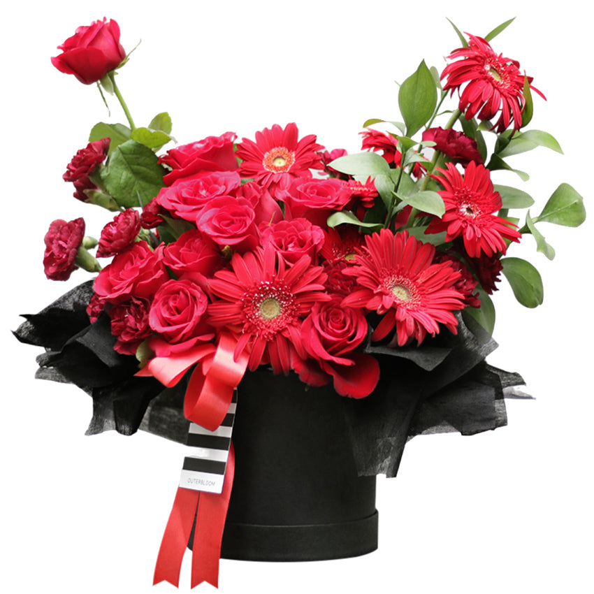 A dramatic and passionate bloom box arrangement overflowing with an assortment of red flowers. It features classic red roses, vibrant red gerberas, and smaller red carnations. The flowers are artfully arranged in a round, matte black box. The arrangement is accented with green leaves and wrapped at the top with ruffled black tissue paper. A striking red satin ribbon and a black-and-white striped ribbon are tied around the box. This bold and romantic gift is perfect for anniversaries and declarations of love