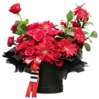 A dramatic and passionate bloom box arrangement overflowing with an assortment of red flowers. It features classic red roses, vibrant red gerberas, and smaller red carnations. The flowers are artfully arranged in a round, matte black box. The arrangement is accented with green leaves and wrapped at the top with ruffled black tissue paper. A striking red satin ribbon and a black-and-white striped ribbon are tied around the box. This bold and romantic gift is perfect for anniversaries and declarations of love