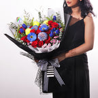 A vibrant and bold bouquet featuring a striking combination of red roses, blue gerberas, and 3 padel/tennis balls. The flowers are arranged with lush green eucalyptus and other filler greens, creating a dynamic contrast. The wrapping is a sophisticated mix of black netted mesh, solid black paper, and a clean white inner layer. The bouquet is tied with a classic black-and-white striped ribbon, adding a modern touch. This powerful arrangement is perfect for celebrating achievements or making a bold state