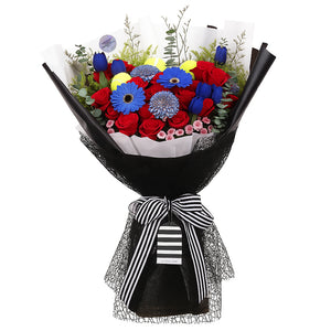 A vibrant and bold bouquet featuring a striking combination of red roses, blue gerberas, and 3 padel/tennis balls. The flowers are arranged with lush green eucalyptus and other filler greens, creating a dynamic contrast. The wrapping is a sophisticated mix of black netted mesh, solid black paper, and a clean white inner layer. The bouquet is tied with a classic black-and-white striped ribbon, adding a modern touch. This powerful arrangement is perfect for celebrating achievements or making a bold state