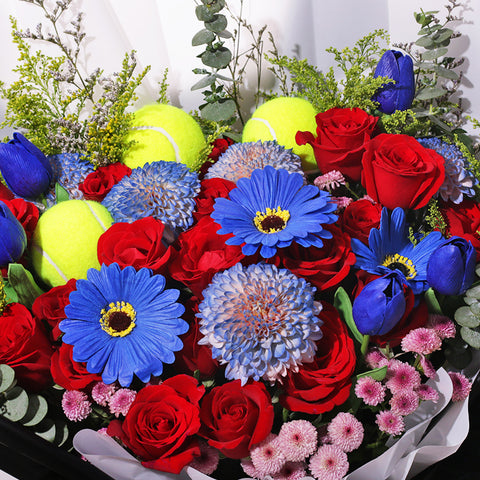 A vibrant and bold bouquet featuring a striking combination of red roses, blue gerberas, and 3 padel/tennis balls. The flowers are arranged with lush green eucalyptus and other filler greens, creating a dynamic contrast. The wrapping is a sophisticated mix of black netted mesh, solid black paper, and a clean white inner layer. The bouquet is tied with a classic black-and-white striped ribbon, adding a modern touch. This powerful arrangement is perfect for celebrating achievements or making a bold state