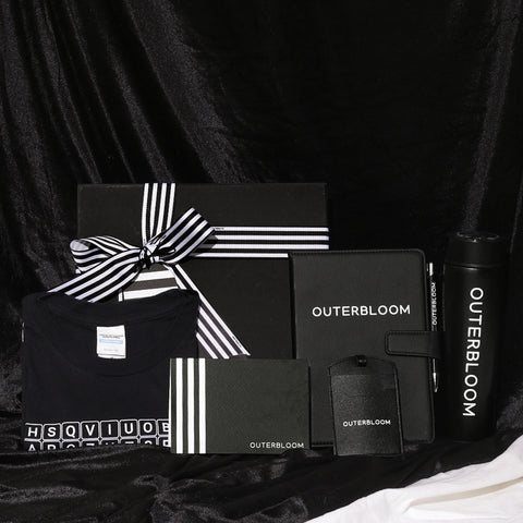 A sophisticated and practical gift hamper with a sleek black and white theme, perfect for corporate gifting or welcoming new team members. The set includes a large black box with a black-and-white striped ribbon bow, a black T-shirt, a black notebook/planner, a black cardholder, and a matching black luggage tag, all featuring the "OUTERBLOOM" logo. A black tumbler is also included, completing this professional and stylish set. The hamper provides a thoughtful collection of useful items for any professional.