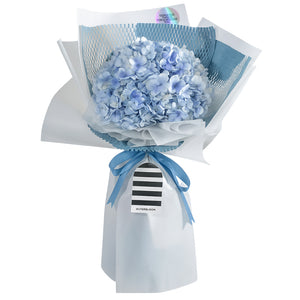 A delicate and elegant bouquet featuring a single, large blue hydrangea head as the centerpiece. The beautiful pastel blue flowers are surrounded by a unique wrapping style: a white honeycomb mesh and a solid baby blue paper, all encased in a soft white outer paper. A light blue satin ribbon is tied in a neat bow, with a stylish black-and-white striped tag attached. A holographic sticker at the top adds a modern, playful touch. This serene bouquet is a gentle and thoughtful gift for any occasion.