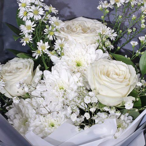 An elegant bouquet featuring a monochromatic white and grey theme. The arrangement is composed of classic white roses, asters, and peacock, delicate baby's breath, and other smaller white filler flowers. The flowers are wrapped in a clean, white inner layer and a sophisticated grey outer layer. The bouquet is tied with a double white ribbon—and a small tag. The minimalist style makes it a thoughtful gift for any occasion.