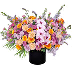 A magnificent flower arrangement bursting with a vibrant mix of colors and textures. This stunning piece features elegant light pink orchids as the centerpiece, surrounded by an array of yellow and orange roses, pink and peach carnations, and various purple and lavender filler flowers like snapdragons. The flowers are arranged in a sleek, round black box. A bold black-and-white striped ribbon is tied into a large bow. This arrangement is a perfect choice for celebrating grand and joyful celebrations.