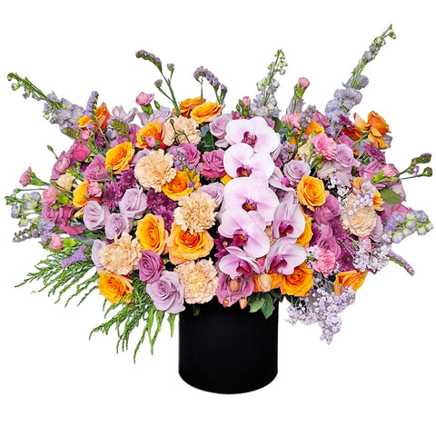 A magnificent flower arrangement bursting with a vibrant mix of colors and textures. This stunning piece features elegant light pink orchids as the centerpiece, surrounded by an array of yellow and orange roses, pink and peach carnations, and various purple and lavender filler flowers like snapdragons. The flowers are arranged in a sleek, round black box. A bold black-and-white striped ribbon is tied into a large bow. This arrangement is a perfect choice for celebrating grand and joyful celebrations.