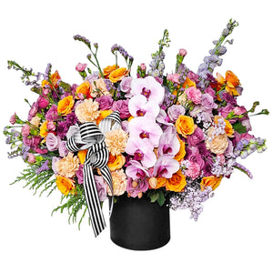 A magnificent flower arrangement bursting with a vibrant mix of colors and textures. This stunning piece features elegant light pink orchids as the centerpiece, surrounded by an array of yellow and orange roses, pink and peach carnations, and various purple and lavender filler flowers like snapdragons. The flowers are arranged in a sleek, round black box. A bold black-and-white striped ribbon is tied into a large bow. This arrangement is a perfect choice for celebrating grand and joyful celebrations.