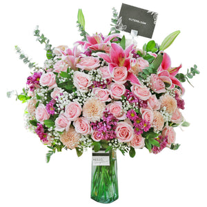 An opulent floral arrangement in a clear glass vase, featuring a magnificent mix of pink and white blooms. The bouquet includes elegant pink lilies, light pink roses, delicate baby's breath, and other pink and purple filler flowers, accented with lush eucalyptus leaves. A striped ribbon and a "Hello gorgeous" card are tied to the vase, with a small black tag at the top. This grand, luxurious display is perfect for celebrating monumental occasions.