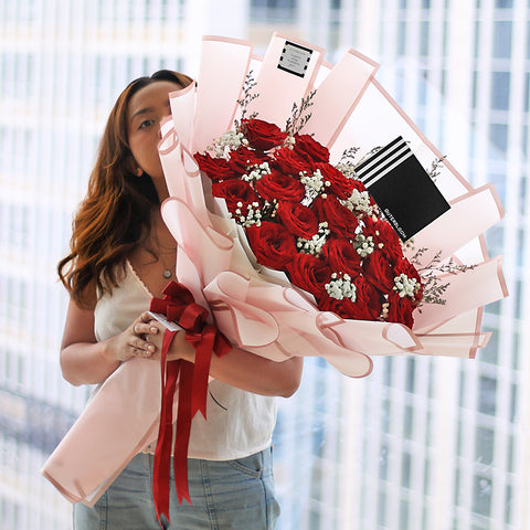 A luxurious and romantic bouquet featuring a generous cluster of approximately 20 red roses. The deep red roses are accented with delicate white baby's breath and thin sprigs of dried filler flowers. The bouquet is wrapped in a soft, semi-transparent pink paper with a subtle striped texture. A double red ribbon is tied into a beautiful bow at the base, and the bouquet includes a stylish black tag. This elegant arrangement is the perfect expression of love and passion.