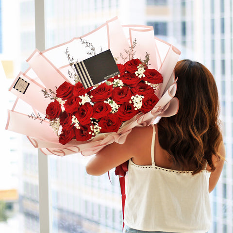 A luxurious and romantic bouquet featuring a generous cluster of approximately 20 red roses. The deep red roses are accented with delicate white baby's breath and thin sprigs of dried filler flowers. The bouquet is wrapped in a soft, semi-transparent pink paper with a subtle striped texture. A double red ribbon is tied into a beautiful bow at the base, and the bouquet includes a stylish black tag. This elegant arrangement is the perfect expression of love and passion.