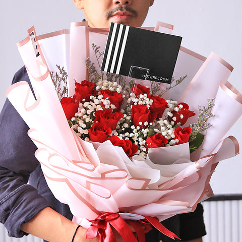 A luxurious and romantic bouquet featuring a generous cluster of red roses. The deep red roses are accented with delicate white baby's breath and thin sprigs of dried filler flowers. The bouquet is wrapped in a soft, semi-transparent pink paper with a subtle striped texture. A double red ribbon is tied into a beautiful bow at the base, and the bouquet includes a stylish black tag. This elegant arrangement is the perfect expression of love and passion.